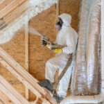 Person fully covered in safety gear, spraying foam insulation in an unfinished attic space.